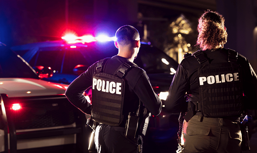 Two police officers in vests with flashing lights in the background, responding to an incident with full kit including secure P25 critical communications radios