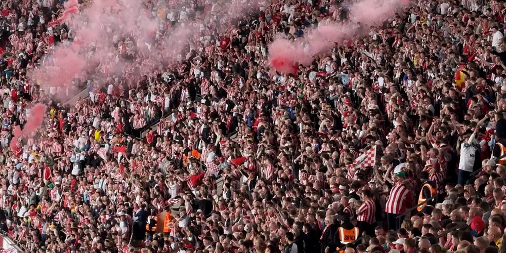 Packed stadium crowd with flags and smoke, showing high-noise, high-density conditions that challenge event communications.