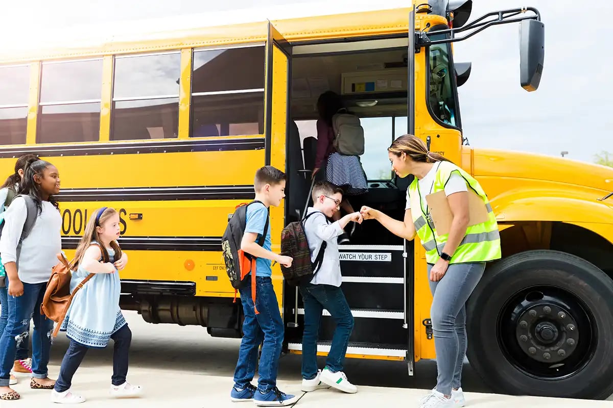Students boarding a school bus while a staff member assists, highlighting the importance of reliable school communications and student safety during transportation.