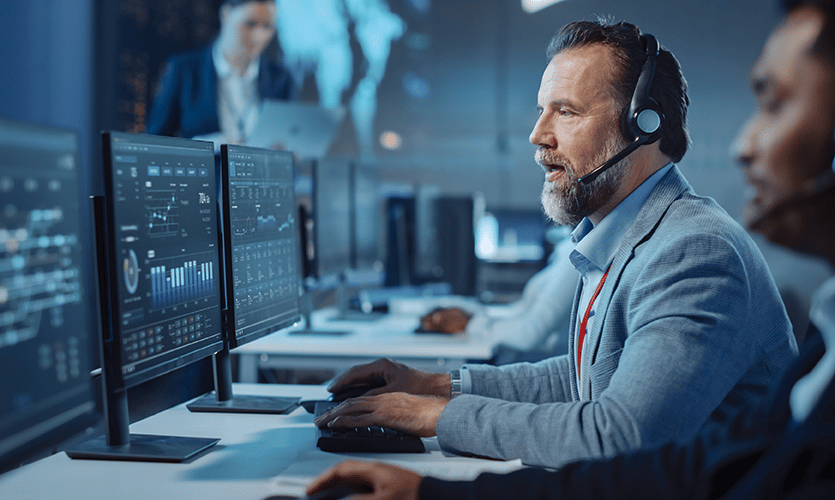 Middle-aged man wearing a headset monitoring a complex network on multiple screens while collaborating with a client.